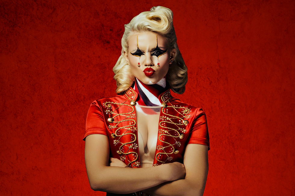 Person with dramatic makeup poses with crossed arms. They wear a red jacket with gold details, set against a deep red background.