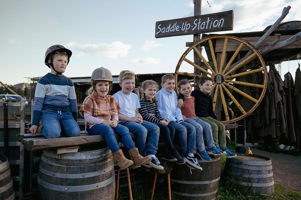 Seven children sit on barrels at a "Saddle-Up Station," wearing helmets and casual outfits, with a wagon wheel and jackets in the background.