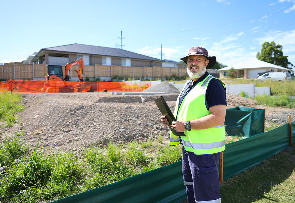 Man in high-vis vest smiles at a construction site with orange fencing, an excavator, and houses in the background under a blue sky.