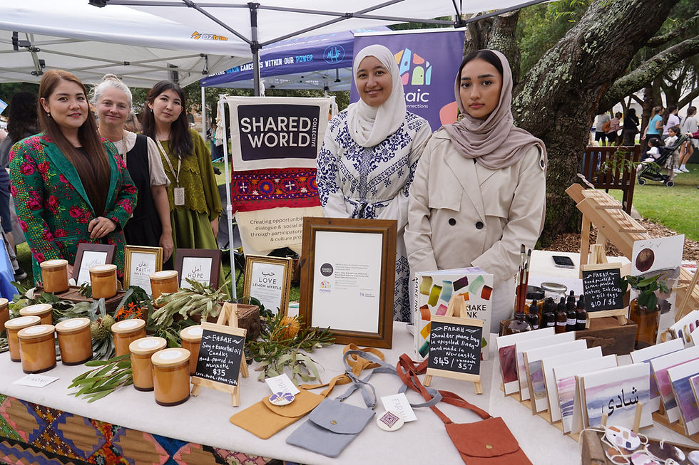 Women in colorful attire stand behind a market stall with handmade candles, bags, and art. Banner reads "Shared World". Outdoor setting.