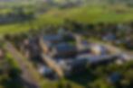 Aerial view of large historic stone building complex with courtyards, surrounded by lush green fields and suburban homes under a clear sky.