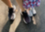 Adult and child feet in sneakers stand on a sunny, textured pavement. Adult wears dark shoes; child wears white with a colorful dress.