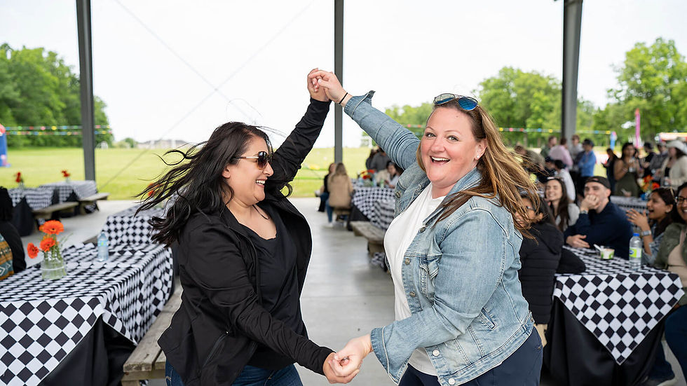 Two women dance happily under a pavilion with checkerboard tablecloths and a green field in the background; a joyful, outdoor gathering.