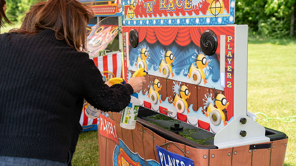 Person playing a "Spray 'N' Race" game with water guns outdoors. Colorful ducks on a carnival booth. Bright, cheerful setting.
