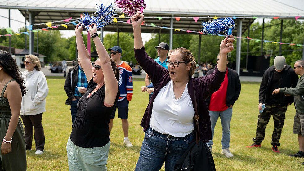 Two women cheer with pom-poms outdoors, surrounded by people. Colorful banners hang above in a park setting on a sunny day.