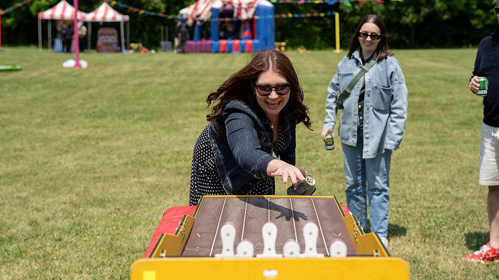 Woman in denim jacket plays carnival game outdoors, laughing. Tent and colorful bunting in background. Friends watch, holding drinks.