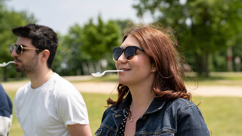 Two people balance spoons with small balls in their mouths outdoors. They're wearing sunglasses and appear focused. Trees and grass in background.