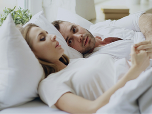 Pessoa pensativa, sentada na cama ao lado do parceiro/parceira que dorme ou lê, sugerindo algo não dito