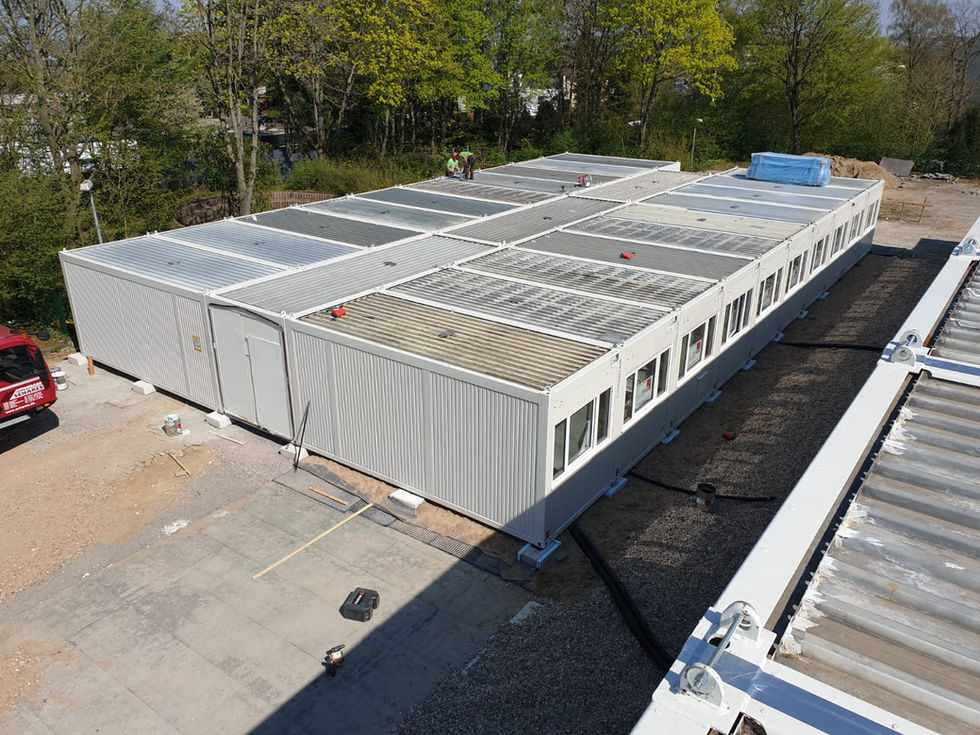 Luftaufnahme einer langen Containeranlage mit Flachdach, während der Bauphase mit Arbeitern auf dem Dach.