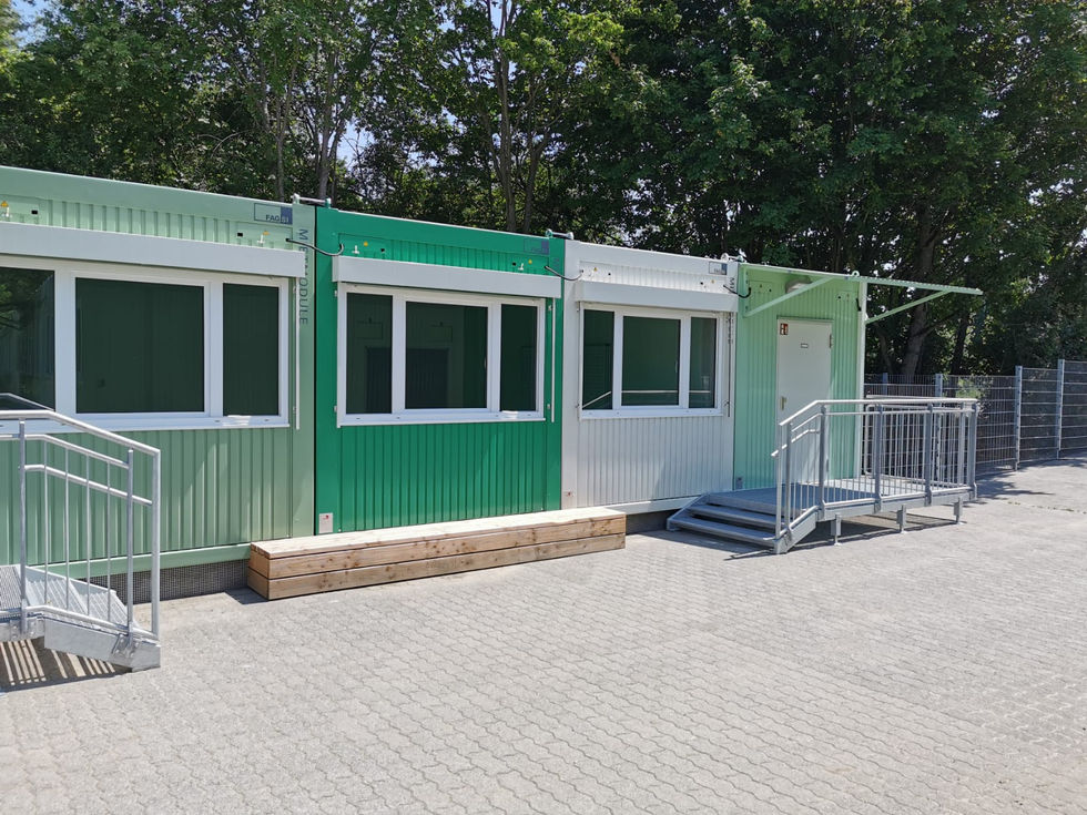 Moderne Container-Schulanlage in Weiß und Grün an einem Hang mit ein- und zweistöckigen Gebäudeteilen, Treppenstufen und Kunstrasen im Außenbereich.