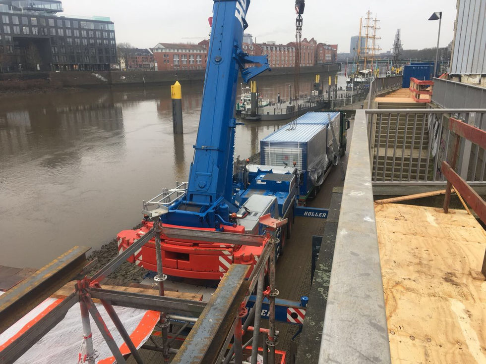 Kran hebt ein blaues Containermodul von einem Lkw auf der Gerüstkonstruktion am Flussufer.