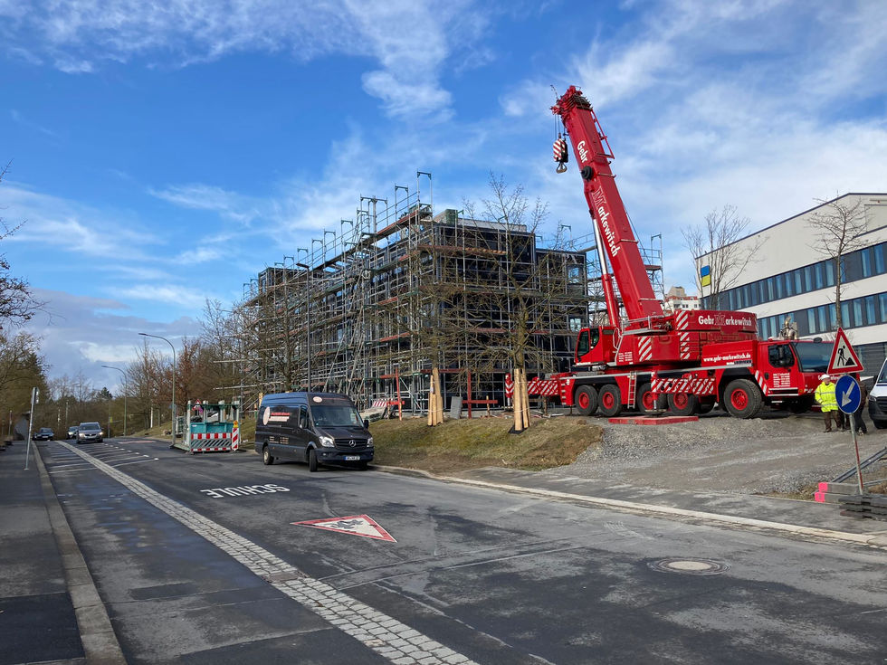 Ein großer roter Mobilkran steht auf einer Schotterfläche neben einer Straße, während im Hintergrund ein mehrstöckiges Modulgebäude für die Montage vollständig mit einem Baugerüst umgeben ist.
