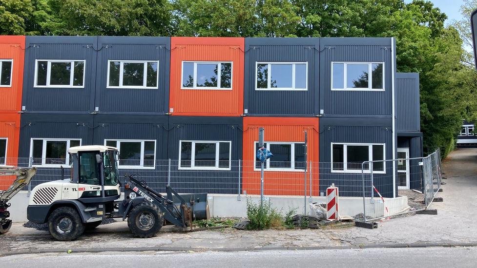 Fertiggestellte Containeranlage in Anthrazit und Orange, davor ein Radlader hinter einem Baustellenzaun.
