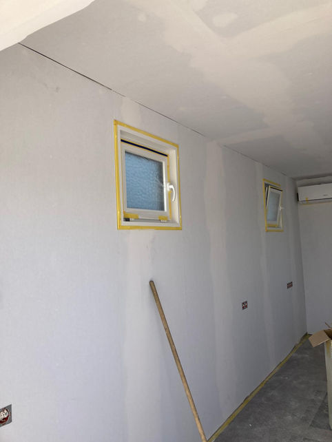 Rohbau-Innenraum mit gespachtelten Wänden, zwei abgeklebten Fenstern, Klimagerät und Sicherungskasten während der Ausbauphase.