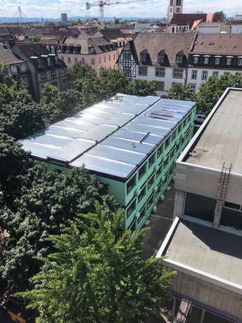 Luftaufnahme eines großen, dreistöckigen Containergebäudes in Hellgrün mit einem glänzenden Blechdach, umgeben von grünen Bäumen und Altstadt-Wohngebäuden unter blauem Himmel.