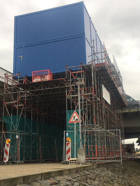 Blauer Containerbau auf massivem Gerüst über einer Brückenunterführung, umgeben von Bauzäunen.