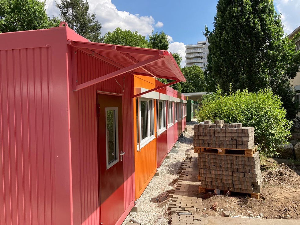 Rot-oranger Containerbau mit Vordach, Eingangstür und Fensterfront, im Hintergrund gestapelte Pflastersteine.