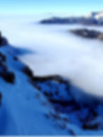 Snowy mountain slope overlooking a valley blanketed in clouds, with distant peaks visible under a clear blue sky. Peaceful and serene.