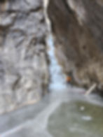 Climber in orange jacket navigates icy terrain in a rocky canyon with frozen waterfall. Cold, adventurous setting with gray and brown tones.