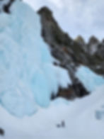 Person climbing ice with gear at base of tall, blue frozen waterfall on snowy mountain. Cloudy sky, rugged terrain, tranquil mood.