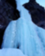 Person ice climbing a frozen waterfall on a cliff. The ice is a light blue, and the climber is secured with a pink rope. Rugged terrain.