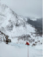 Climber in red jacket ascends icy slope; snowy mountains and cloudy sky in background. Rugged, cold, adventurous setting.