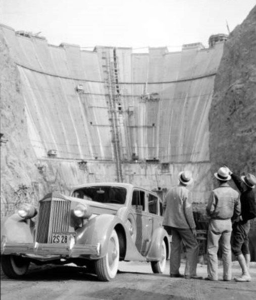 Vintage car and three men in hats admire a massive dam under construction; steep walls, scaffolding visible. Mood is awe and curiosity.