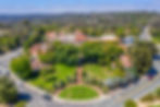 Aerial view of a green park with trees surrounded by buildings with red roofs. Roads with cars, stop and arrow signs are visible.