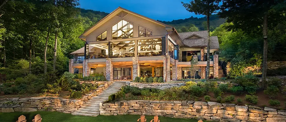 Large, modern house with stone accents, surrounded by lush trees and gardens. Outdoor lights illuminate the scene, creating a serene evening mood.