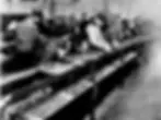 Factory workers assembling products on a long conveyor belt. The setting is industrial with metallic parts and machinery. Mood is focused.