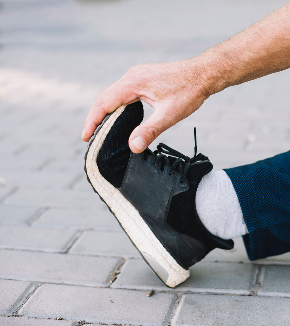 Hand stretching a leg, touching a black sneaker with white sole on a paved surface. Casual setting, blue pants, neutral mood.