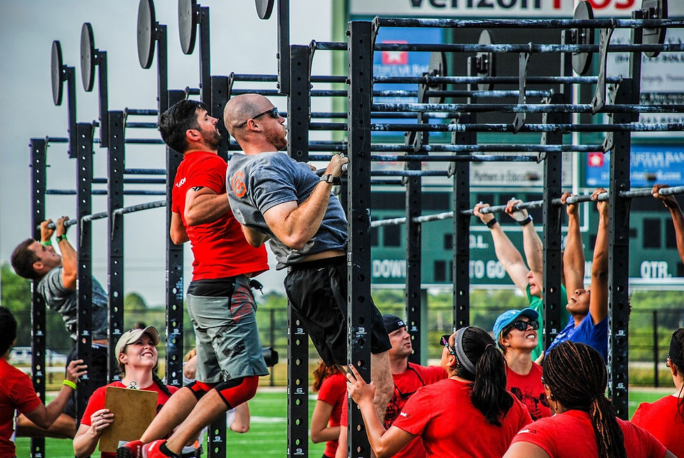 People exercising outdoors on pull-up bars; some wear red shirts. A coach with a clipboard watches. Bright day, stadium backdrop.