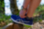 Person ties blue running shoe on a wooden railing in a leafy area, wearing a watch. Bright sunlight enhances the colors.