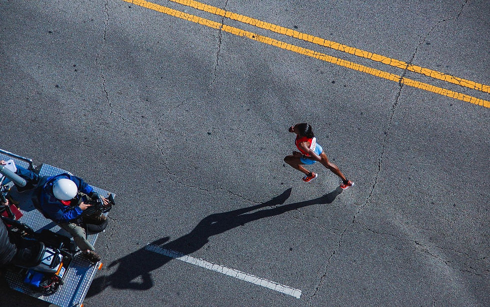 Runners in colorful shoes and sportswear race on a sunlit street. Bib numbers visible. Energetic scene with movement and focus on legs.