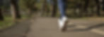 A person in blue pants jogs on a path through a park with trees, wearing white sneakers. It's a sunny, peaceful day.