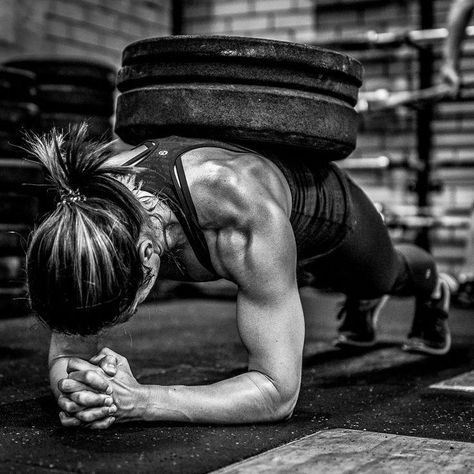 A woman performs a plank with weights on her back in a gym. She's focused, showing strength. Black and white image, brick wall backdrop.