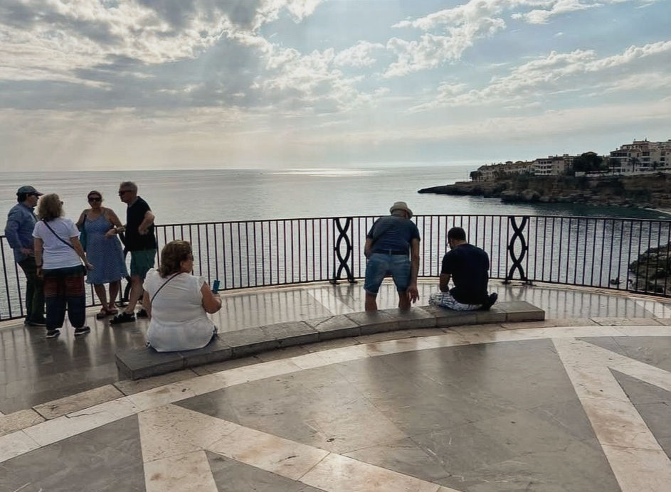 People on a terrace overlooking the sea, some sitting, some standing. Cloudy sky, distant town on a cliff, relaxed atmosphere.