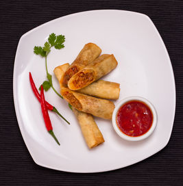 Food stylist photographer Dubai captures crispy spring rolls with chili dip on white plate, styled with red peppers and cilantro on dark background