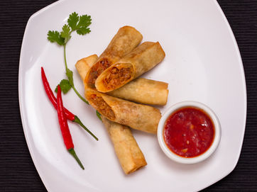 Food stylist photographer Dubai captures crispy spring rolls with chili dip on white plate, styled with red peppers and cilantro on dark background