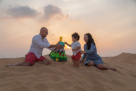 Documentary family photography of parents and toddler decorating a small Christmas tree on desert dunes at sunset.
