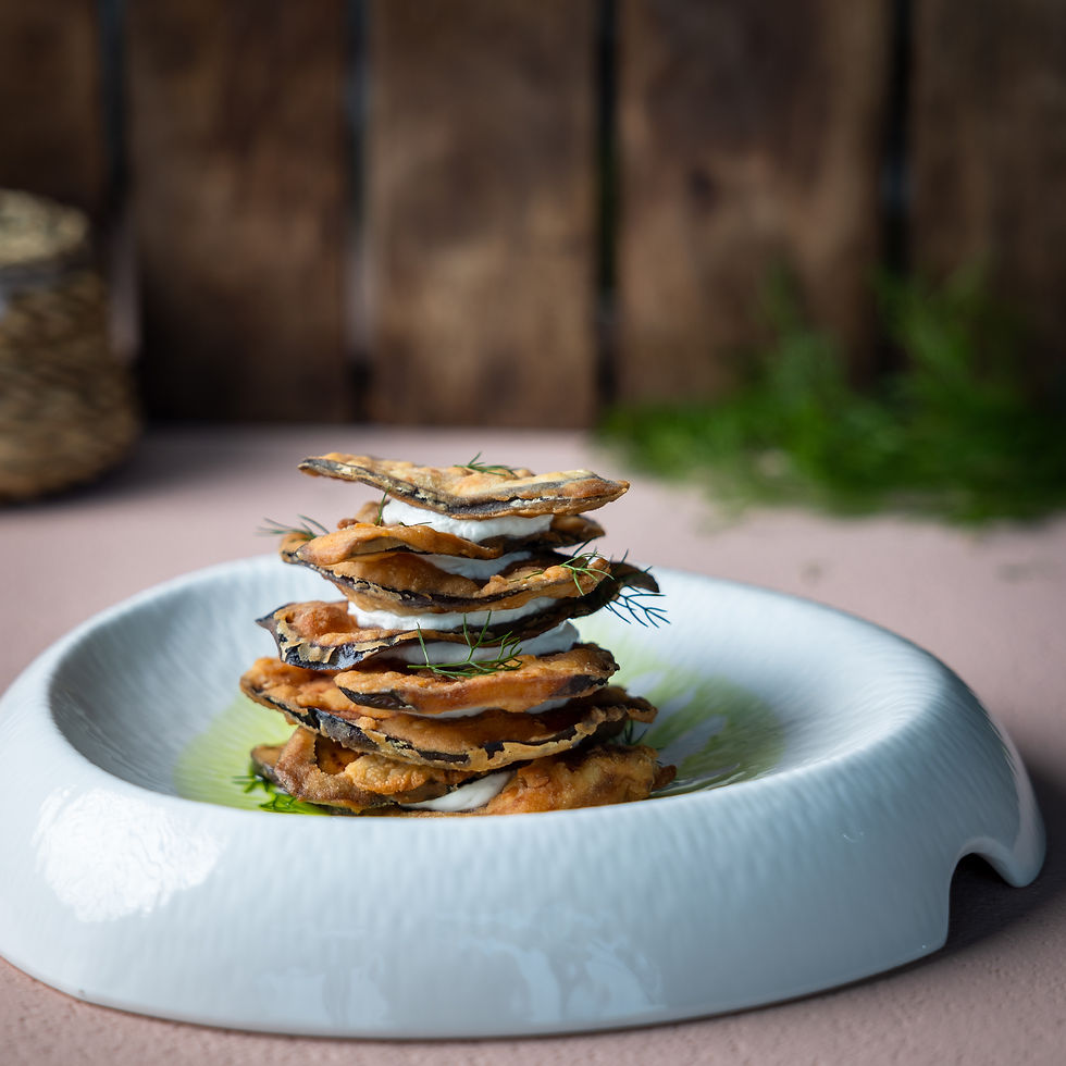 Close-up food photography of a crispy eggplant stack with creamy layers and dill on a white bowl, rustic wood background, and soft light.