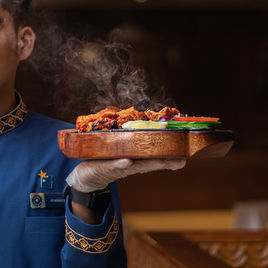 Food photographer shot of a server in blue uniform presenting a sizzling wooden platter with steam, grilled kebabs, and fresh vegetables.