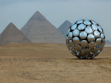 product photography: reflective spherical sculpture in desert with Giza pyramids background, metallic mirror finish, dramatic overcast sky