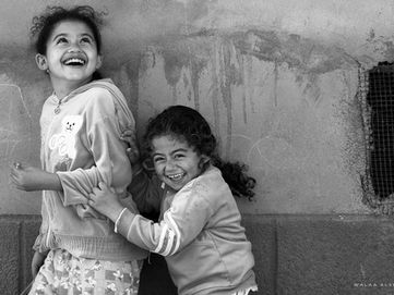 Documentary photo, black-and-white of two laughing girls playing against a rough wall—candid expressions, joy, and innocence.