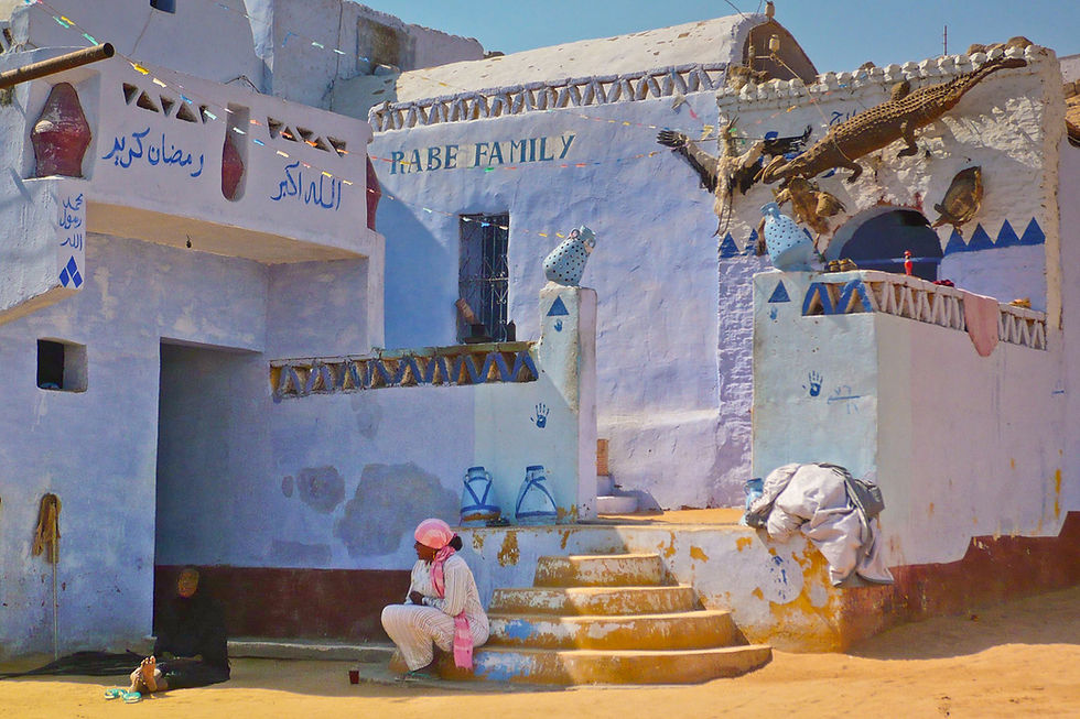Documentary photo of a blue-painted Nubian house with crocodile ornaments; a man on the steps and a woman by the doorway in bright daylight.