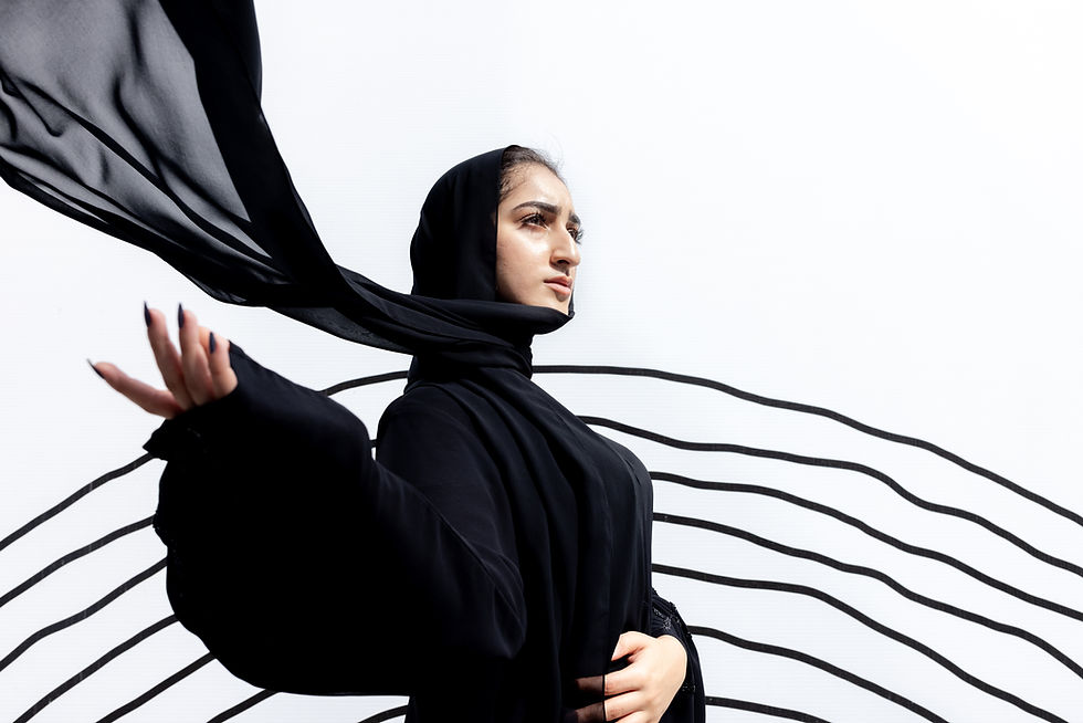 Documentary Portrait of an Arab woman in a flowing abaya, scarf in motion against a minimal white wall with bold black lines.