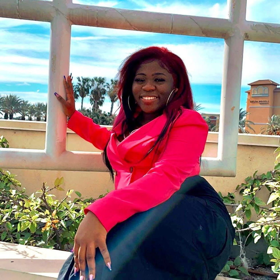 Smiling woman in a pink blazer poses by a window frame, under a clear blue sky with palm trees and a hotel in the background.