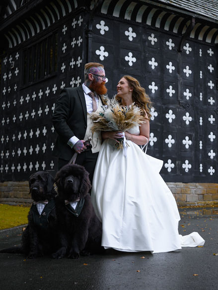 Alternative wedding photography of a joyful couple with their dogs, standing by a historic black-and-white house in a non-traditional setting.