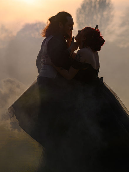Unique alternative wedding photography of a couple in dark attire sharing an intimate moment amidst mist and a soft glowing sunset.