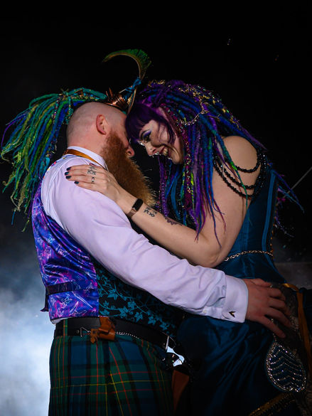 Gothic wedding couple with vibrant, fantasy-inspired attire share a tender embrace in front of a train under a dramatic night sky.
